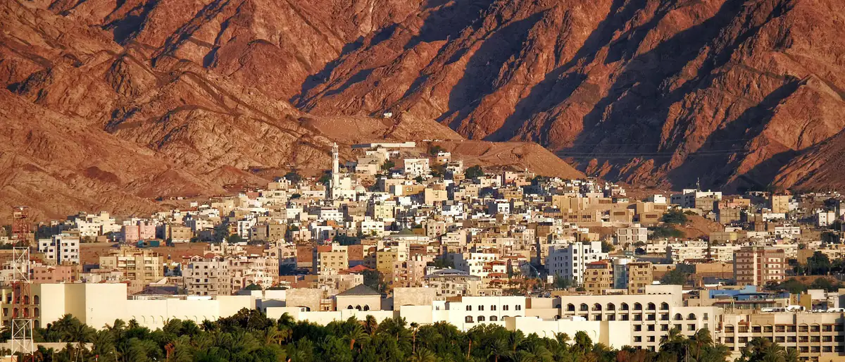 مواقيت الصلاة في العقبة: مدينة العقبة بين جبالها الحمراء وأشجار النخيل، وتظهر مئذنة مسجد بين البيوت