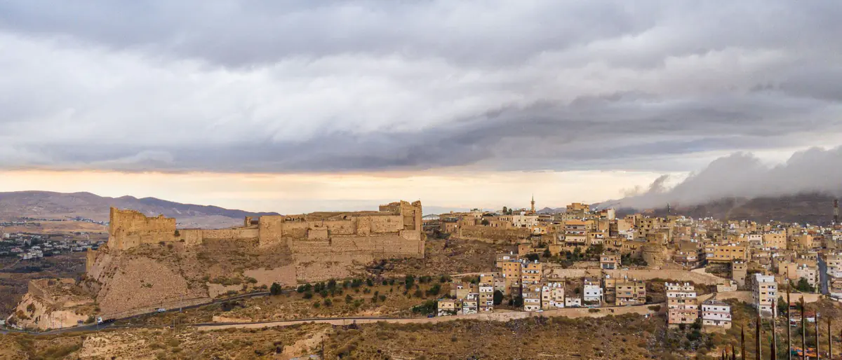 مواقيت الصلاة في الكرك: قلعة الكرك على حافة الجبل وإلى جانبها بيوت مدينة الكرك ومئذنة، تحت غيوم رمادية
