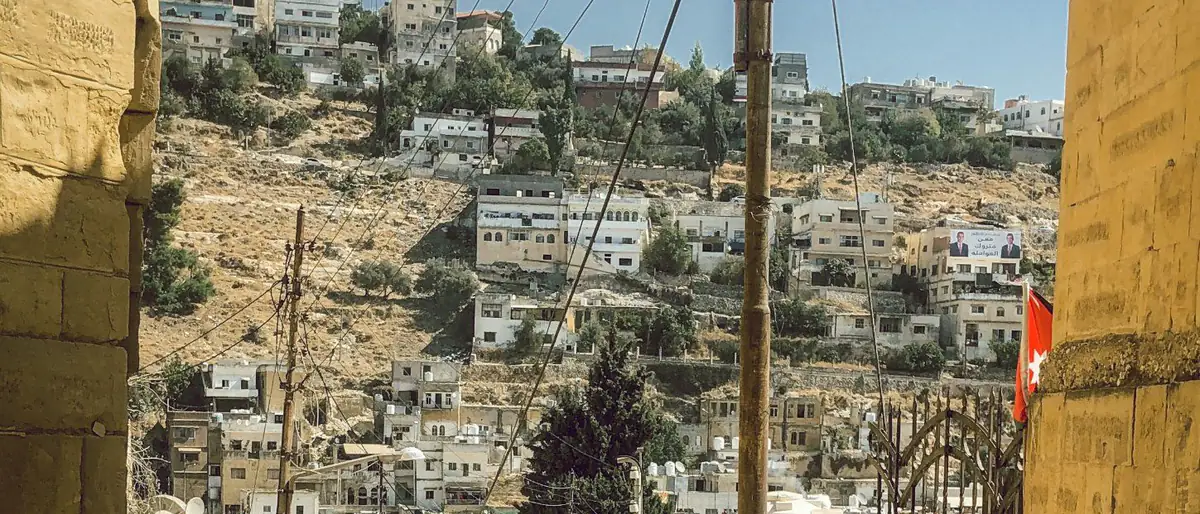 مواقيت الصلاة السلط: بيوت على سفح جبل تظهر بين جدارين من الحجر الأصفر في حي قديم، وعلم الأردن على أحد البيوت
